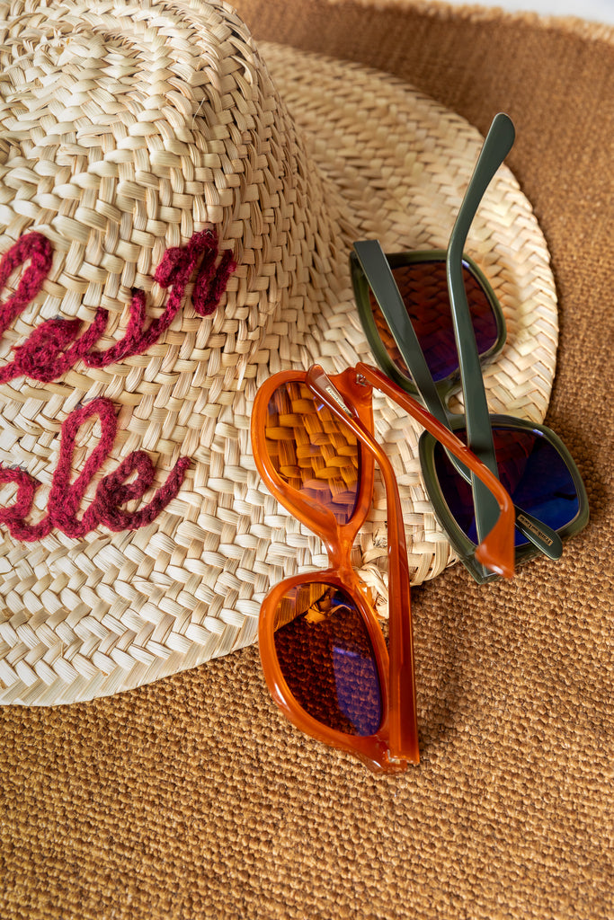Summer flatlay with straw hat embroidered with “Couleur Locale” and two pairs of Frank and Lucie Eyewill sunglasses – one in orange, one in green, placed on woven mat